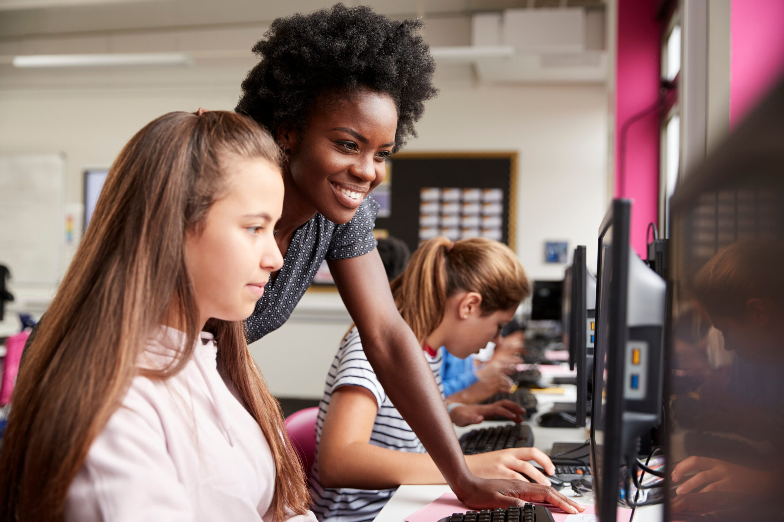 Teacher Helping Female Pupil Line Of High School Students Working at Screens In Computer Class